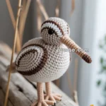 A crocheted snipe bird with brown and white patterns, featuring black eyes and a long beak, stands on a wooden surface near a yellow leaf.