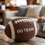 A knitted football-shaped pillow with the words 'GO TEAM' sits on a wooden table in a cozy living room setting.