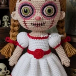A crocheted doll with braided hair, large green eyes, and stitched details, wearing a white dress with red trim, resembling a spooky or Halloween-themed character.