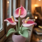 A crocheted plant featuring pink flowers and green leaves in a crocheted pot, placed on a wooden windowsill in a cozy room.