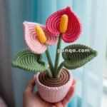 A crochet calla lily plant with pink and red petals and yellow spadices, held in a hand in front of a blue curtain.