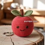 A crocheted red apple with a smiling face and two green leaves on top, attached to a keyring, placed on a wooden surface.