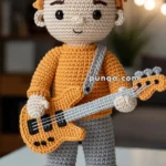 A crocheted doll with orange hair styled in spikes, dressed in an orange sweater and gray pants, holding a crocheted orange guitar with a gray strap. The doll is wearing orange and white sneakers and stands on a white surface with a blurred background.