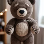 A crocheted teddy bear with brown and beige yarn, standing on a wooden surface with a small sprig of lavender in front of it. The background is softly lit with warm fairy lights.