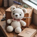 A crocheted teddy bear keychain with a pink bow tie sits on a wooden surface beside a wooden box and a small booklet titled "Dream Big."