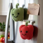 Two crocheted bell peppers, one green and one red, with smiley faces are attached to a stainless steel refrigerator door. A note reading "FRESH" is taped nearby.