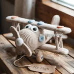 A crocheted toy biplane with large expressive eyes sitting on a wooden table near a window, featuring two wings adorned with orange and blue details. The table holds a tag with the phrase 'fly high.'
