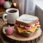 Crocheted sandwich with layers of colorful yarn resembling lettuce, cheese, and meat between two slices of bread, alongside a crocheted cup of coffee and a strawberry, all placed on a wooden surface.