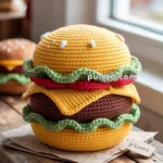 A crocheted hamburger plushie featuring a yellow bun with sesame seeds, green lettuce, red tomato, cheese, and a brown patty, placed on a wooden table beside a paper with "yum" written on it.