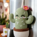 A crocheted green cactus with a pink flower on top, featuring smiling eyes, hangs from a refrigerator in a kitchen. There's a green mug labeled 'COFFEE' on the fridge and a blurred background with plants and kitchen decor.