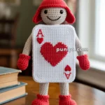 A crocheted doll wearing a red hat and red gloves, designed to resemble an Ace of Hearts playing card with a smiling face, standing on a wooden surface with blurred background.