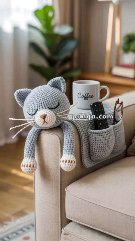 A crocheted gray cat with closed eyes is draped over the arm of a beige sofa, next to a mug labeled "Coffee" and an armrest pocket holding a remote control and glasses.