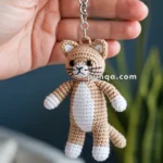 A hand holds a crocheted cat keychain with a beige and white body, featuring black button eyes and embroidered facial details.