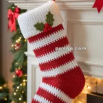 A festive red and white crocheted Christmas stocking with a holly leaf and berries decoration is hanging from a decorated mantel with garland, lights, and ornaments.