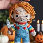 A hand holding a crocheted doll keychain with orange hair, wearing a rainbow-striped sweater and blue overalls, and holding a small knife, set against a background with pumpkins and candles.