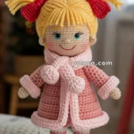 A crocheted doll with yellow yarn hair styled in pigtails tied with red bows, wearing a pink coat, scarf, and boots, set against a cozy indoor background with blurred Christmas decorations.