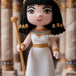 A crocheted doll dressed in ancient Egyptian attire, featuring a white gown, a colorful beaded collar, and a gold crown with a serpent motif. The doll is holding a golden staff and is standing in a setting with Egyptian-style columns.