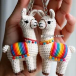 A hand holding two crocheted llama keychains, each adorned with colorful rainbow blankets and decorative accents, connected to a metal key ring.