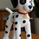 Crocheted dalmatian dog with black spots and a red collar with a yellow tag, positioned on a wooden surface.