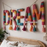 A bedroom features a decorative wall hanging that spells "DREAM" in colorful, knitted letters with tassels. The letters are hanging above a bed with a rattan headboard, adorned with beige pillows and a textured throw pillow. A plant is positioned to the side of the bed.