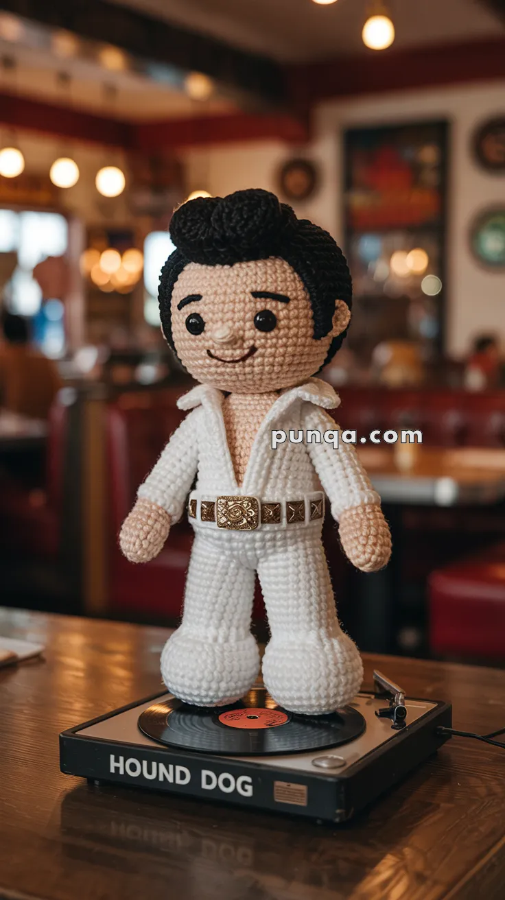 Crocheted Elvis Presley doll dressed in a white jumpsuit standing on a record player labeled 'Hound Dog' in a retro-style diner setting.