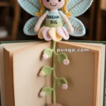 A crocheted fairy with wings, wearing a "Dream Big" dress, adorns a bookmark inside an open book. The bookmark features a green crochet stem with pink flowers and a mushroom at the bottom.