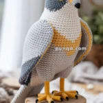 A crocheted falcon, resembling a peregrine falcon, is perched on a wooden stump with a tag reading 'Handmade with love.' The bird features detailed knitted textures, including gray, white, and yellow sections, with black accents around the eyes and on the beak and feet.