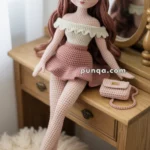 A crocheted doll with long pink hair and a white off-shoulder top, sitting on a wooden dresser near a mirror, with a matching pink skirt and a small pink purse beside it.