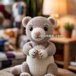 A crocheted toy ferret with a light brown and cream color scheme is sitting on a round wooden table in a cozy room with blurred background featuring a lamp and plants.