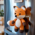 A crocheted fox toy attached to the front of a stainless steel fridge, with a small wooden sign reading "Welcome" hanging above it.