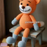 A crocheted fox toy with orange body, gray feet and hands, and a smiling face sits on a small bench surrounded by small crochet books in various colors.