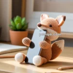 A crocheted fox-shaped phone holder on a wooden desk, with a smartphone held between its paws, and a potted plant in the background.