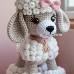 A crocheted plush toy poodle with beige and white yarn, featuring a decorative pink bow and collar with a gold medal. The background shows a soft pink chair.