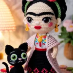 A crocheted doll with colorful flowers in its hair and intricate embroidered clothing stands on a pink doily with a small crocheted black cat beside it.