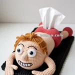 A crocheted tissue box cover shaped like a cartoon character lying on its stomach, featuring a humorous facial expression with large eyes and an exaggerated smile.