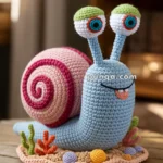 A crocheted snail with a blue body, large white eyes with green tops, and a pink and red spiral shell sits on a wooden table. It is adorned with colorful crocheted elements resembling plants around its base.