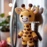 A crocheted giraffe magnet with brown spots and a smiling face attached to a stainless steel refrigerator. In the background, there's a blurred view of kitchen decor and a red apple on a wooden countertop.