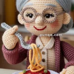A crocheted figure of an elderly woman with glasses and gray hair, smiling while holding a fork with yarn spaghetti over a yarn plate of spaghetti with tomato sauce and peas.