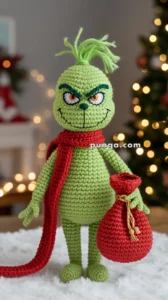 A crocheted green character with a mischievous smile, wearing a red scarf and holding a red sack, against a festive, blurred background with lights.