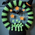 A Halloween-themed crochet wreath featuring a black and green striped design with a black witch hat, orange stars, and a bat. A crochet cauldron with bubbling potion hangs at the bottom.