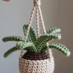 A hand holding a small crocheted potted plant with green leaves and a brown base, suspended by a looped cord. The plant is made with yarn and has a decorative, handmade appearance.