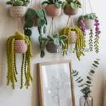 Crocheted hanging planters with green leaves and colorful stems displayed on a wooden shelf, next to a framed botanical print and a small amber handbag.