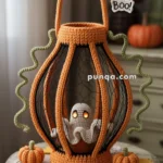 A crocheted Halloween decoration featuring a ghost inside a cage-like structure with orange and black yarn. The ghost has glowing eyes, and the decoration is set on a small table with mini crocheted pumpkins and a "Boo!" tag hanging above.