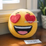 A crocheted yellow emoji pillow with heart-shaped eyes and a smiling expression sits on a wooden surface next to a small note that says 'You're Loved.'