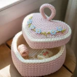 A heart-shaped pink crocheted basket with a lid decorated with blue flower embroidery, containing sewing thread and small sewing accessories, placed on a wooden stool by a window.
