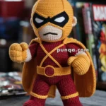 A crocheted doll resembling a superhero figure, wearing a yellow and red costume with a cape, black mask, and belt, posed with fists raised. The background features blurred comic books on a shelf.
