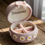 A crocheted jewelry box adorned with small white and yellow flowers, containing rings and labeled "Treasures" inside the lid, placed on a wooden table.
