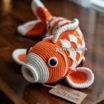 A crocheted orange and white fish toy on a wooden table with a tag reading "Handmade with love."
