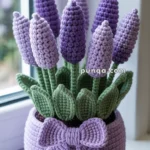A crochet potted plant featuring purple lavender-like flowers with green leaves and a lilac pot adorned with a matching bow, placed on a window ledge.