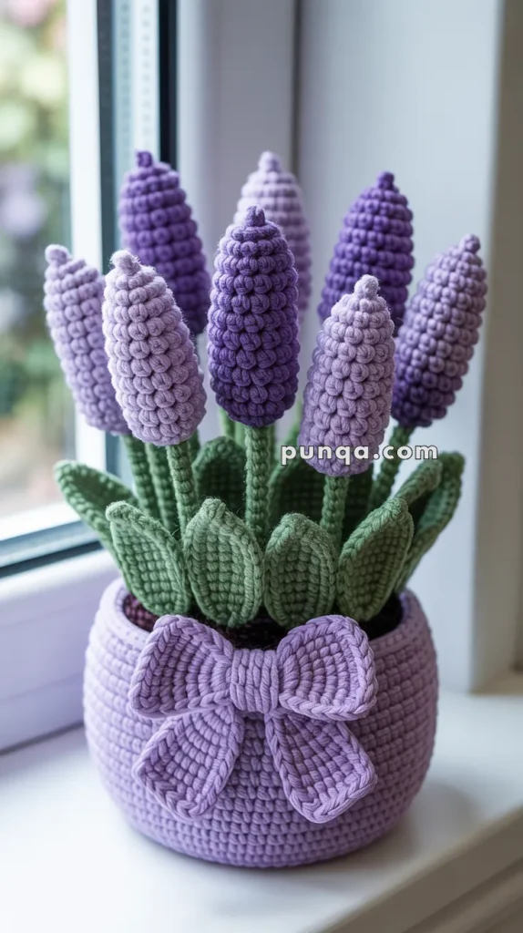 A crochet potted plant featuring purple lavender-like flowers with green leaves and a lilac pot adorned with a matching bow, placed on a window ledge.